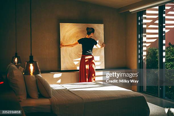 woman adjusting large abstract canvas on wall inside sunlit bedroom. wearing rust-colored pants and black top. interior styling moment. - vida sencilla fotografías e imágenes de stock