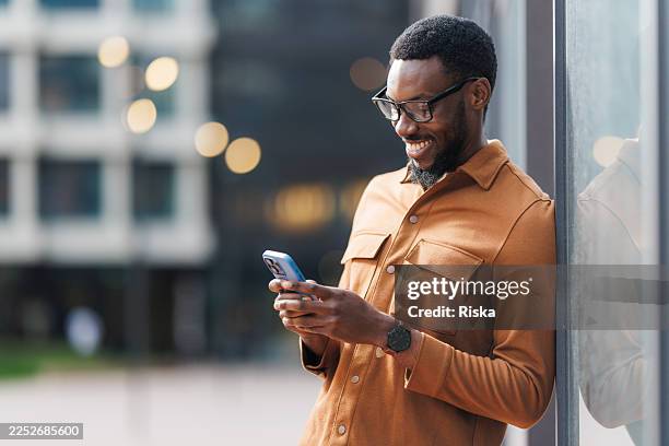 young man standing outdoors, smiling and using smartphone - modern manhood stock pictures, royalty-free photos & images