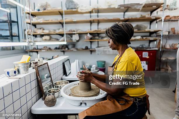 senior woman modeling clay on a pottery wheel in an art studio - kunstatelier stockfoto's en -beelden