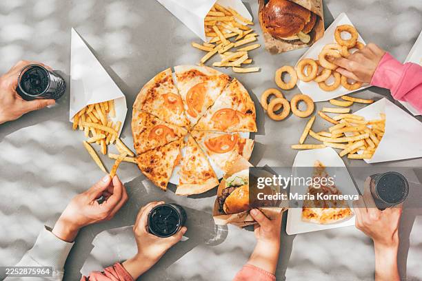 an overhead flat-lay showing multiple hands reaching for pizza, burgers, fries, and onion rings on a grey table. this vibrant social scene captures the concepts of shared meals, casual dining, and togetherness. - fast food stock pictures, royalty-free photos & images