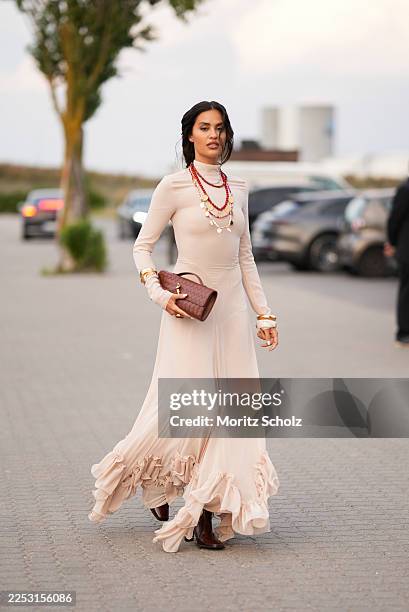 Jessica De Oliveira is seen wearing a long-sleeved, high-neck nude maxi dress with a fitted bodice and flowing skirt with dramatic ruffled hem...