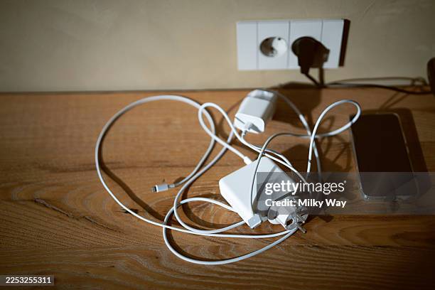 minimalist wooden table with tangled white charging cables, phone and adapters next to a wall socket in warm side lighting. - charging cable stock pictures, royalty-free photos & images