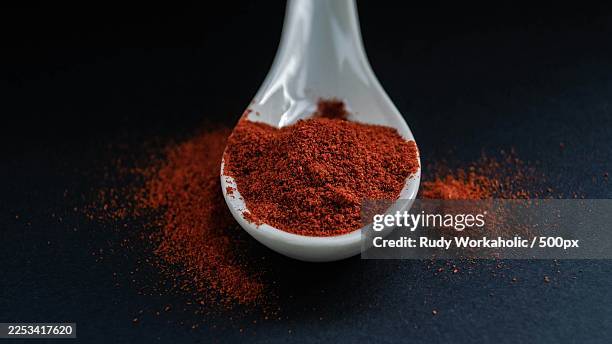 close-up of vibrant red ground chili pepper in a white spoon on a dark background, with scattered spice - paprika stock pictures, royalty-free photos & images