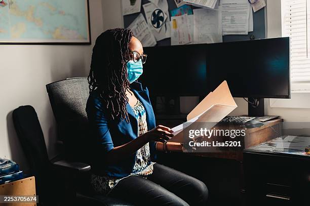 african american woman wearing a mask at work in a home or professional office setting looking at file folders documents and healthcare or tax paperwork - compromised immune system stock pictures, royalty-free photos & images