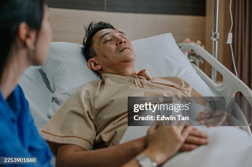 Asiatischer männlicher Patient ruht im Krankenhausbett, während seine Frau seine Hand hält Asiatischer männlicher Patient ruht im Krankenhausbett, während seine Frau seine Hand hält