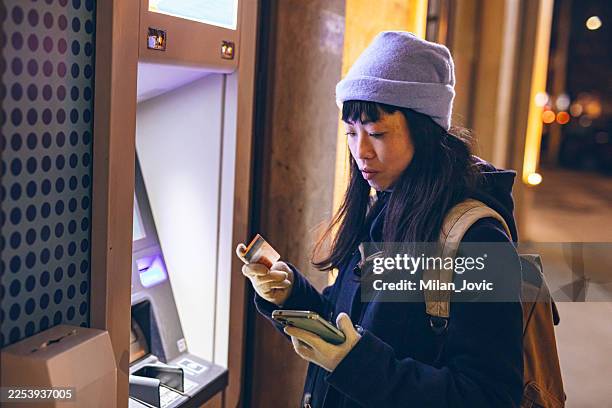 woman using atm while checking smartphone - log on stock pictures, royalty-free photos & images