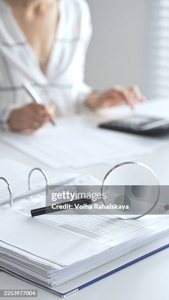 Magnifying glass on financial documents is in focus while an accountant using a calculator and taking notes, emphasizing accounting, finance, close up vertical view. Audit and taxes