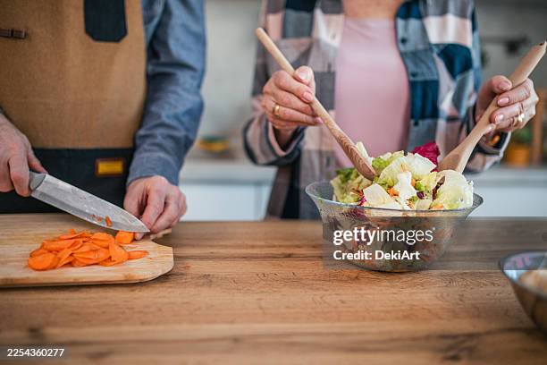 close-up of the hands of a senior man and a woman preparing lunch at the kitchen counter, making a salad and chopping vegetables - salad bowl stock pictures, royalty-free photos & images