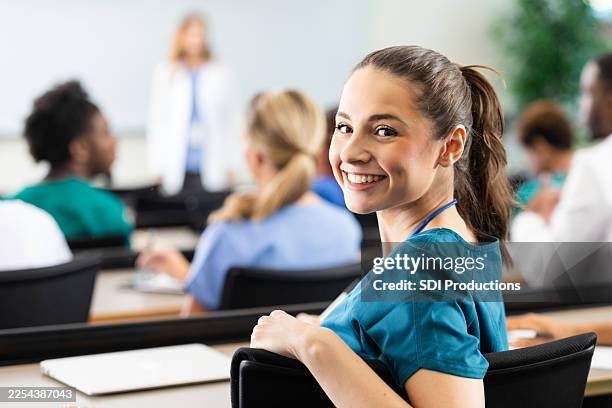 smiling young woman in scrubs in a classroom setting with others present - estudante de medicina imagens e fotografias de stock