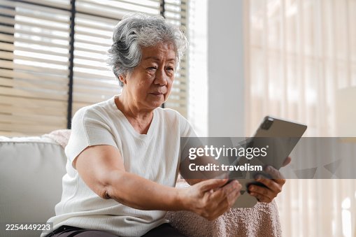 An elderly Asian woman with gray hair relaxes peacefully on a sofa at home, using a tablet at a comfortable reading distance without glasses, enjoying a warm, quiet moment of rest, comfort, and digital connection in a cozy home lifestyle An elderly Asian woman with gray hair relaxes peacefully on a sofa at home, using a tablet at a comfortable reading distance without glasses, enjoying a warm, quiet moment of rest, comfort, and digital connection in a cozy home lifestyle