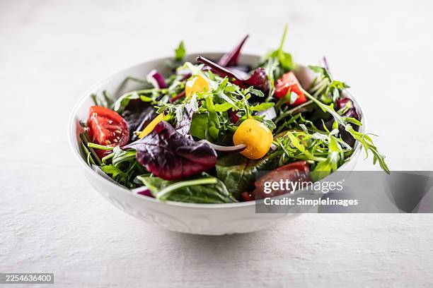 a white bowl full of fresh and colorful salad, featuring leafy greens such as arugula and baby spinach, topped with chunks of ripe red and yellow tomatoes and red onion. - salad bowl stock pictures, royalty-free photos & images