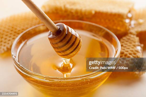 close-up of a honey dipper dripping golden honey into a glass bowl with honeycomb in the background - honing stockfoto's en -beelden