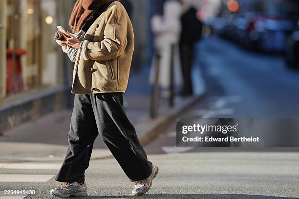 Passerby wears a brown scarf, a brown shearling aviator jacket, black wide leg pants, Asics silver and gray sneakers shoes, holds a mobile phone,...