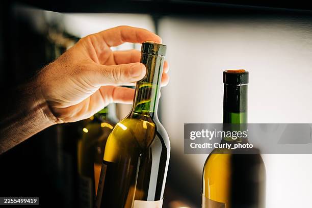 a close-up of a person's hands carefully selecting a bottle of white wine from a minimalist, backlit rack. the warm illumination and human touch evoke a sense of expertise and curation. - witte wijn stockfoto's en -beelden