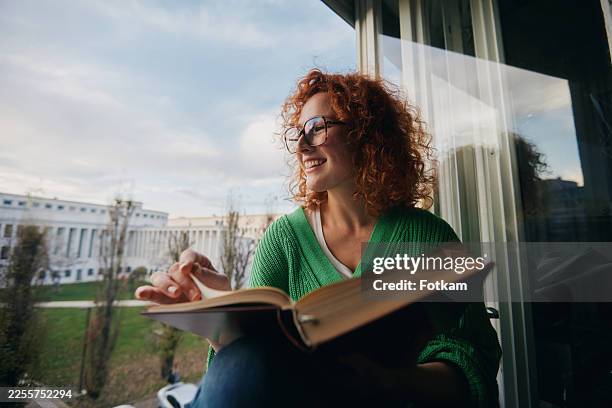 una donna adulta dai capelli rossi legge un libro seduta sul bordo di una finestra - occhiali da lettura foto e immagini stock