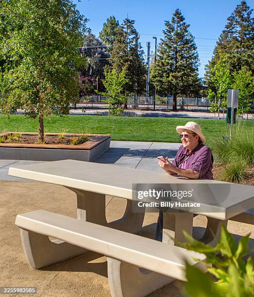 senior citizen holding a cellphone while sitting alone at a new and clean concrete picnic bench in a public park - hårda material landskapsarkitektur bildbanksfoton och bilder
