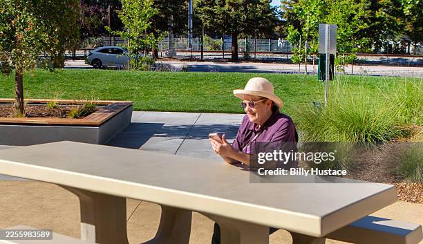 smiling senior citizen holds a cellphone while sitting alone at a new and clean concrete picnic bench in a public park - hårda material landskapsarkitektur bildbanksfoton och bilder
