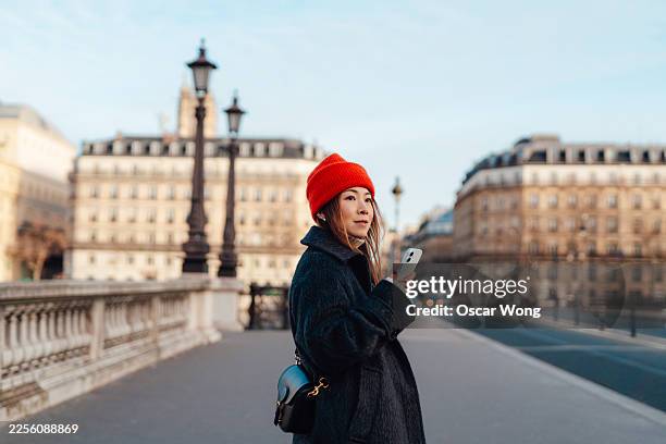 young asian woman standing on a street in paris, using her smartphone - virtual assistant stock pictures, royalty-free photos & images