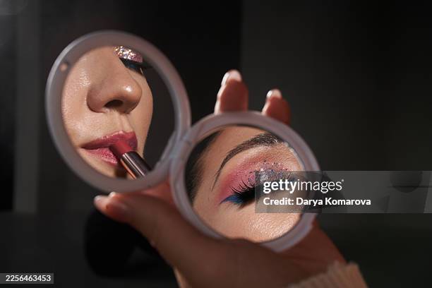 close-up of lipstick application and glitter eye makeup reflected in a compact mirror held by hands - maquillaje de teatro fotografías e imágenes de stock