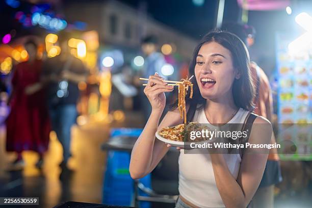 young woman enjoying street food noodles in night market - thai food stock pictures, royalty-free photos & images