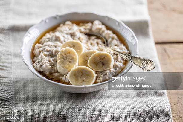 a close-up of a bowl of creamy oatmeal drizzled with maple syrup and garnished with banana slices, served on a linen tablecloth. - harina de avena fotografías e imágenes de stock