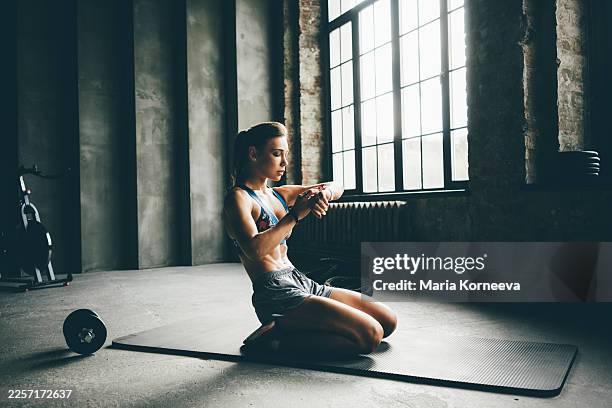 a fit woman sits on a yoga mat in a moody loft gym, checking her smartwatch during a break. - slank bildbanksfoton och bilder