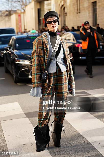 Guest wears a black pillbox hat, black sunglasses, brown plaid blazer with green and orange accents over a grey vest and a black turtleneck, matching...