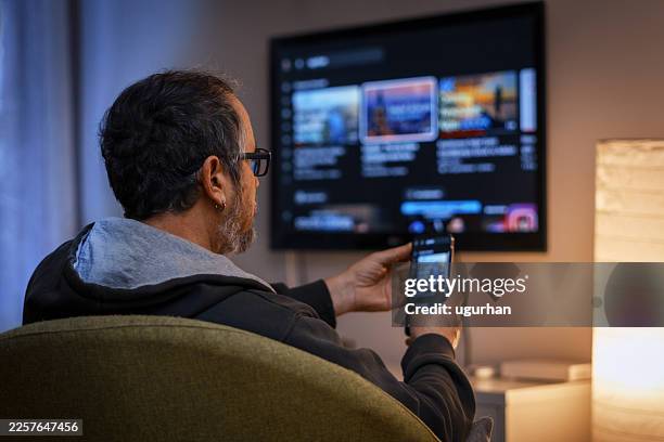 mature man relaxing watching smart tv at home using phone app - smart tv stock pictures, royalty-free photos & images