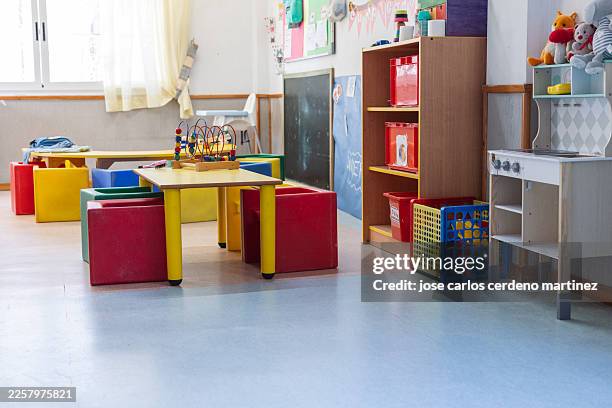 empty classroom in a brightly lit preschool with small wooden tables, yellow chairs, and colorful wall decorations, emphasizing early childhood education - escuela preescolar fotografías e imágenes de stock