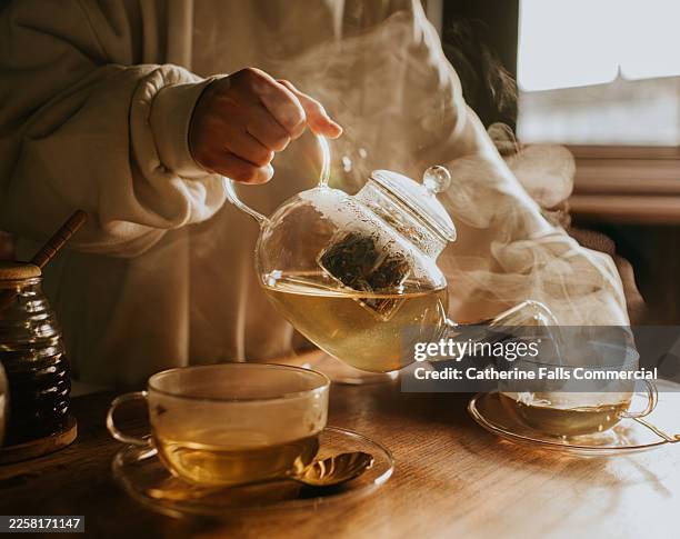 person pouring green tea from a glass teapot into a clear cup by a window. - tea room stock pictures, royalty-free photos & images