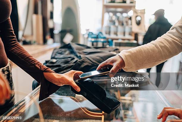 hand of man using tap to pay method through smart phone at checkout counter in clothing store - contactless payment stock pictures, royalty-free photos & images
