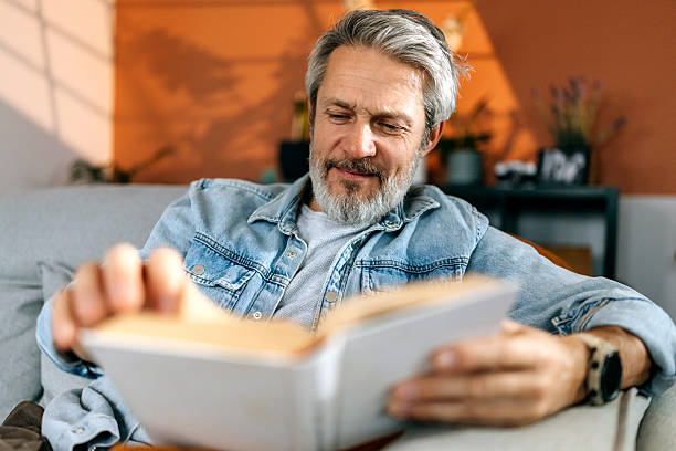 smiling senior man reading book at home - books stock pictures, royalty-free photos & images