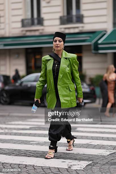 Sara Coelho Ansari wears black hair covered with a black structured headwrap hat, metal drop earrings, metal necklaces with a pendant, a handheld...