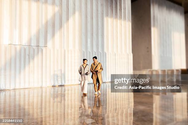 two confident asian businesspeople talking and walking enter a large, light filled interior space together after working. it could be a commercial building or a hotel or luxury apartments. they look around in awe. - plano geral imagens e fotografias de stock