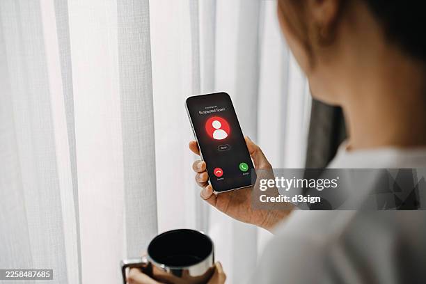 young asian woman receiving an incoming suspected call from unknown caller on her smartphone and rejecting the call at home. device screen showing suspected scam as detected by network provider. phone scam. cyber security and fraud concept - wirtschaftskriminalität stock-fotos und bilder