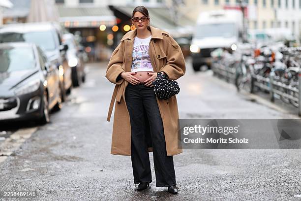 Mary L. Jean is seen wearing a camel colored long wool coat with a trench coat silhouette, epuallette shoulders and a clean button front from Ganni;...
