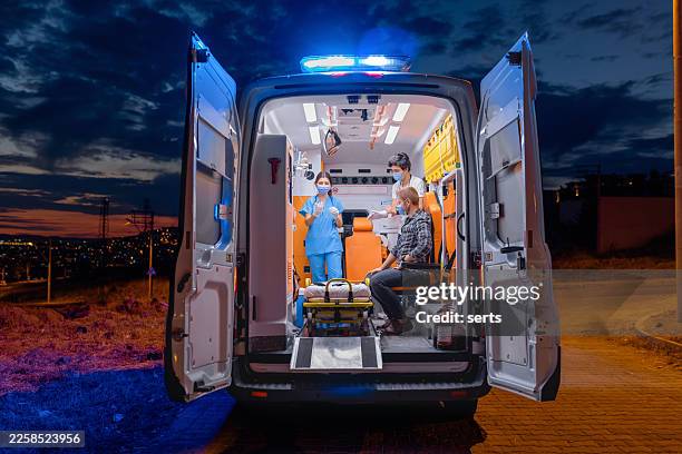 équipe médicale d’urgence assurant des soins de nuit à l’intérieur d’une ambulance ouverte avec des lumières clignotantes et une silhouette urbaine - véhicule de service durgence photos et images de collection