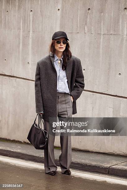 Annabel Rosendahl wears a charcoal grey oversized blazer, a blue striped button-down shirt, grey tailored trousers, a black baseball cap, round...