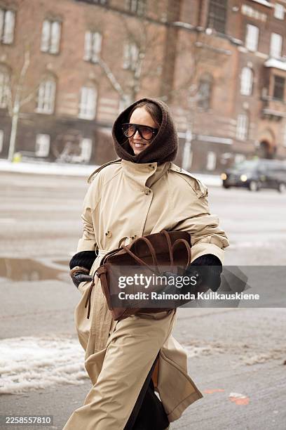Guest wears a beige trench coat cinched at the waist with a matching belt, black leather gloves, oversized brown scarf wrapped around the head and...