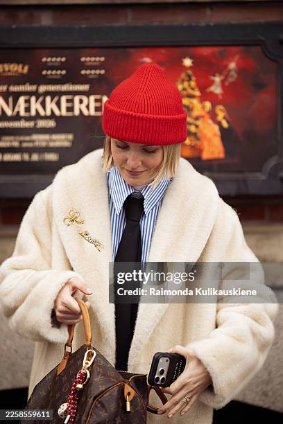 Polly Newman wears a cream faux fur coat, a blue striped button-up shirt, a black tie, black tailored trousers, black loafers, a red knit beanie,...