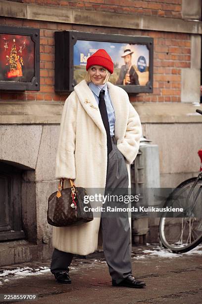 Polly Newman wears a cream faux fur coat, a blue striped button-up shirt, a black tie, black tailored trousers, black loafers, a red knit beanie,...