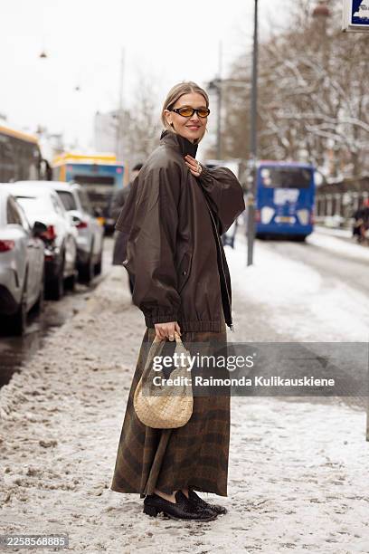Hollie Mercedes Peters wears a brown oversized bomber jacket, a plaid midi skirt, black textured shoes, yellow-tinted sunglasses, and carries a woven...