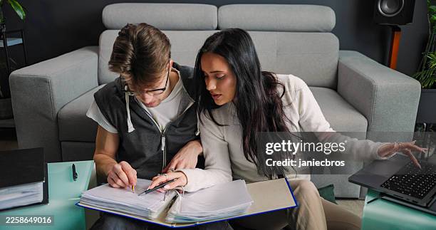 young couple filling out tax documents at home, husband assisting his wife with completing paperwork, seated on the floor beside sofa in a modern living room, pointing out invoice - hot desking stock pictures, royalty-free photos & images