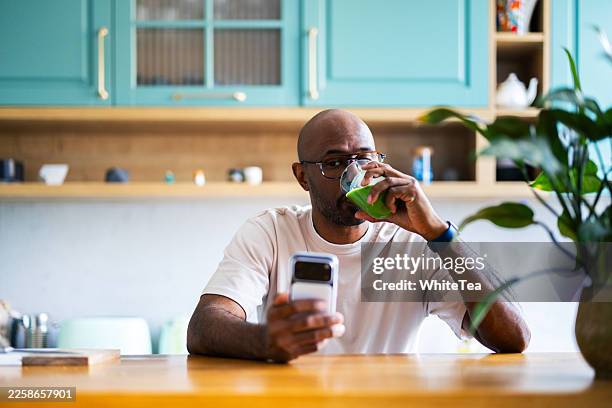 man drinking green juice while browsing phone in kitchen - wheatgrass drink stock pictures, royalty-free photos & images