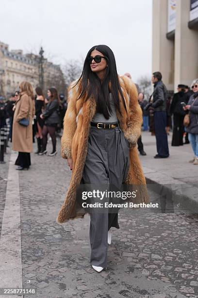 Guest wears long straight black hair with a center part, black rectangular sunglasses, a brown tan fur coat with a wide collar and long length, a...