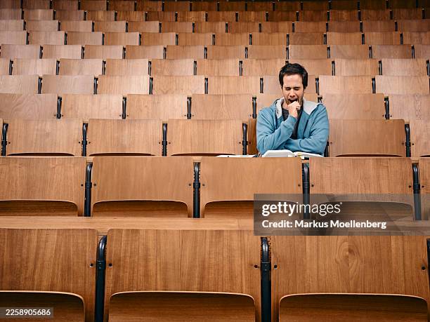 man in a blue sweater sitting and studying in a large empty wooden lecture hall. germany - coursework stock pictures, royalty-free photos & images