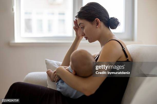 tired young mother holding newborn baby and touching forehead on sofa - postpartum depression stock pictures, royalty-free photos & images