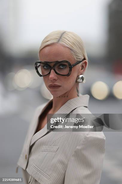 Ava Black seen wearing oversized black retro glasses, silver bold statement earrings, a full ivory pinstriped Karpova Ukraine suit. Her hair is...