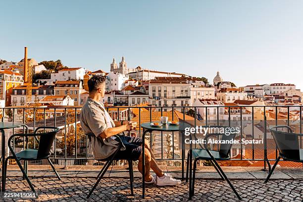 man drinking coffee at a sidewalk cafe with view of lisbon cityscape on a sunny summer day, portugal - wonderlust imagens e fotografias de stock