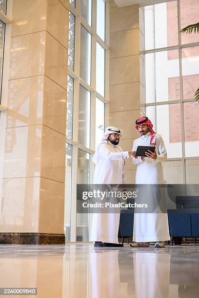 full length shot of two saudi business men in thawbs discussing with tablet in modern hotel lobby interior - arab culture stock pictures, royalty-free photos & images